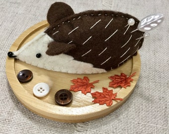 以下が含まれることがあります： 茶色と白のフェルトでできたハリネズミの針山で、葉っぱと真珠のアクセントが付いています。針山は木製の台座の上に置かれており、オレンジ色のフェルトのメープルリーフが3枚とボタンが2つ付いています。