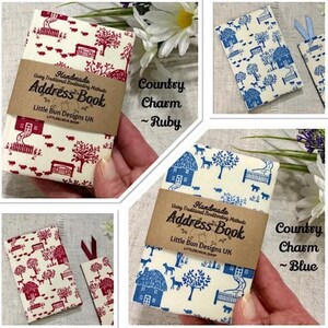 May include: Address books with country charm patterns. One has a ruby red design of houses, trees, and animals on cream. Another has a blue design. Each book has a brown paper band with the text "Address Book".