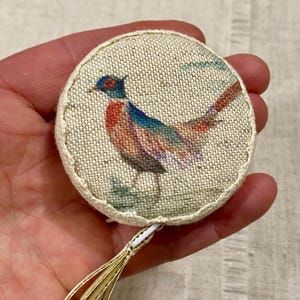 May include: A round, beige fabric measuring tape with a gold tassel. The tape features a colorful pheasant illustration.