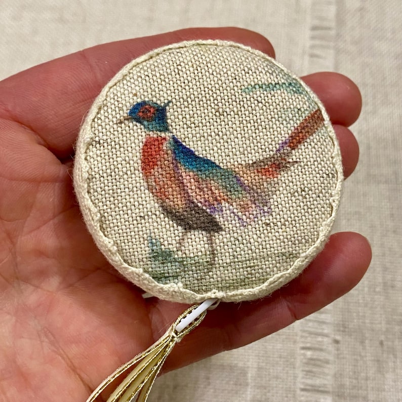 May include: A round, beige fabric measuring tape with a gold tassel. The tape features a colorful pheasant illustration.