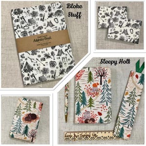 May include: A collection of camping-themed stationery items. Includes an address book, two small notebooks, a pen, a bookmark, and a notebook with a forest design. The address book is wrapped with a brown band and has the text "Hand Covered Address Book".