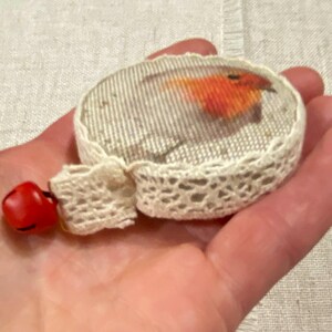 May include: A small, round, white fabric ornament with a robin bird image inside. The ornament is decorated with white lace and has a red bell attached.