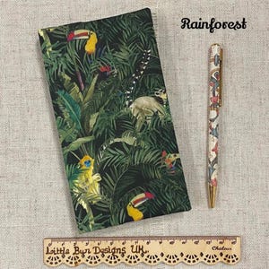 May include: A notebook and pen set with a rainforest theme. The notebook features a dark green cover with toucans, monkeys, and lush foliage. The pen has a white background with colorful floral and animal designs. The word "Rainforest" is written in cursive.