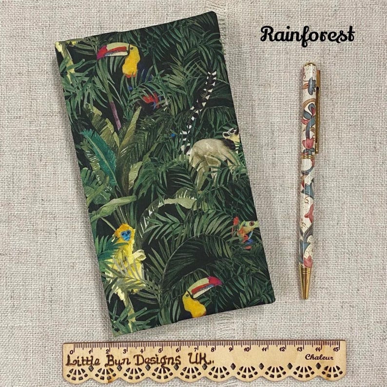 May include: A notebook and pen set with a rainforest theme. The notebook features a dark green cover with toucans, monkeys, and lush foliage. The pen has a white background with colorful floral and animal designs. The word "Rainforest" is written in cursive.