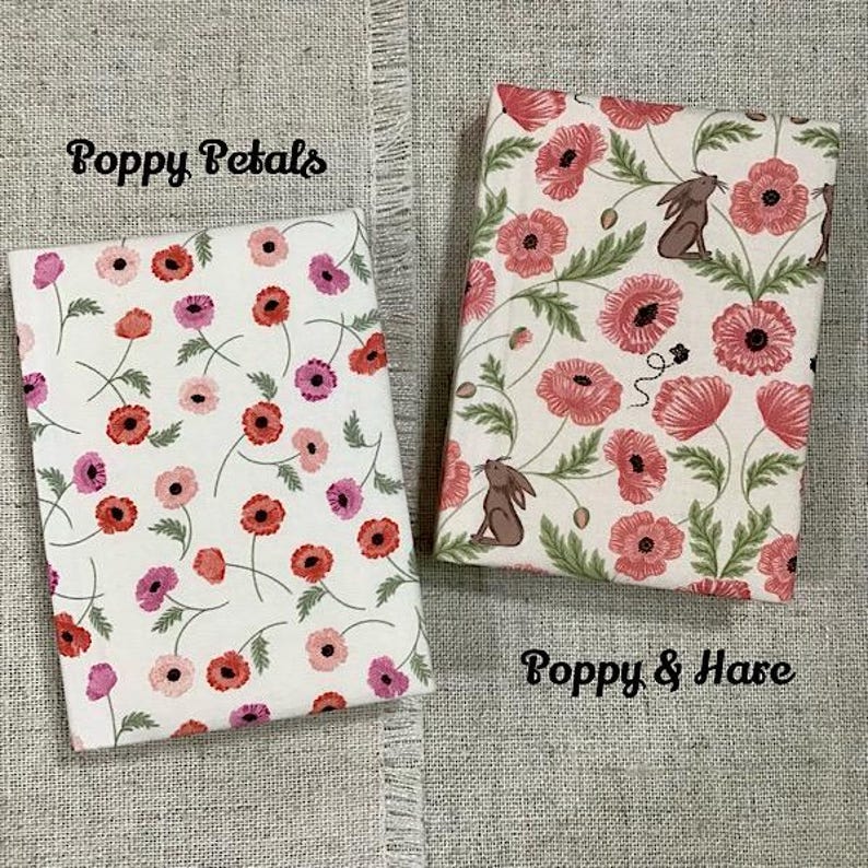 May include: Two fabric-covered books with floral patterns. One features red and pink poppies with the text "Poppy Petals." The other has poppies, a bunny, and the text "Poppy & Hare." Both are on a neutral linen surface.