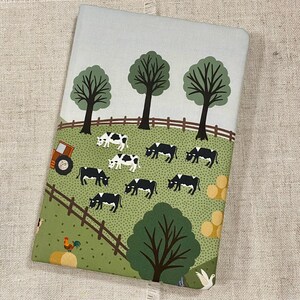 May include: A green and blue illustrated notebook cover with a farm scene. The cover features a tractor, cows, trees, and a fence.