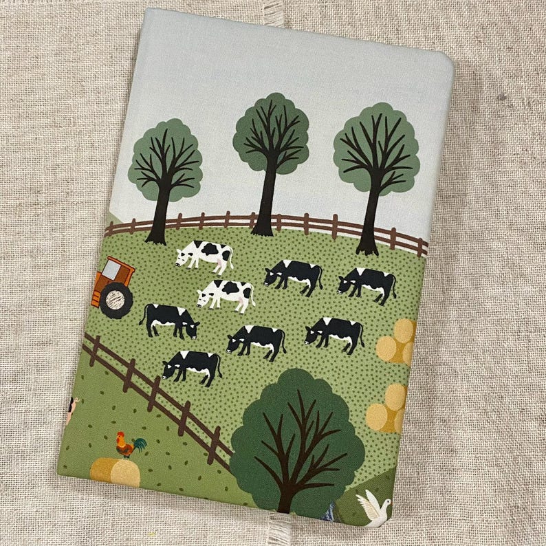 May include: A green and blue illustrated notebook cover with a farm scene. The cover features a tractor, cows, trees, and a fence.