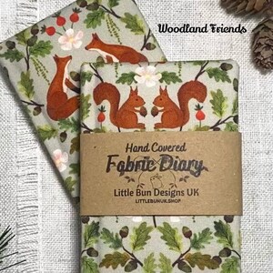 May include: Two fabric-covered diaries with a woodland theme. The fabric features squirrels, foxes, flowers, and foliage in shades of brown, green, and pink. The diaries are wrapped with a brown paper band that reads "Hand Covered Fabric Diary."