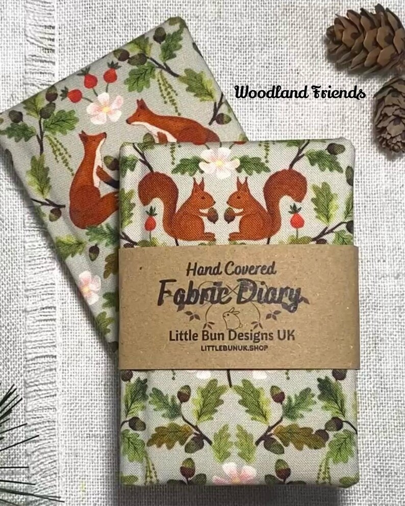 May include: Two fabric-covered diaries with a woodland theme. The fabric features squirrels, foxes, flowers, and foliage in shades of brown, green, and pink. The diaries are wrapped with a brown paper band that reads "Hand Covered Fabric Diary."