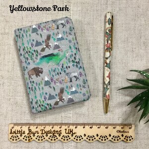 Op de afbeelding: Een grijs notitieboek met stoffen kaft en een Yellowstone Park-print met bergen, bomen, adelaars en bizons, vergezeld van een pen met bloemmotief en een houten liniaal van 15 centimeter lang.