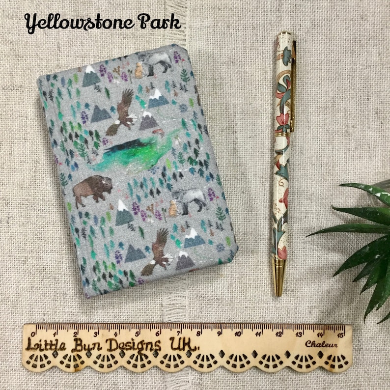 Op de afbeelding: Een grijs notitieboek met stoffen kaft en een Yellowstone Park-print met bergen, bomen, adelaars en bizons, vergezeld van een pen met bloemmotief en een houten liniaal van 15 centimeter lang.