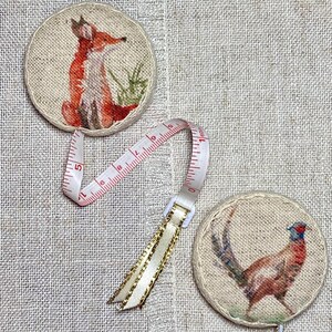 May include: Two round fabric measuring tapes with a fox and a pheasant design. The tapes are beige with brown stitching and feature a white plastic measuring tape with red numbers.
