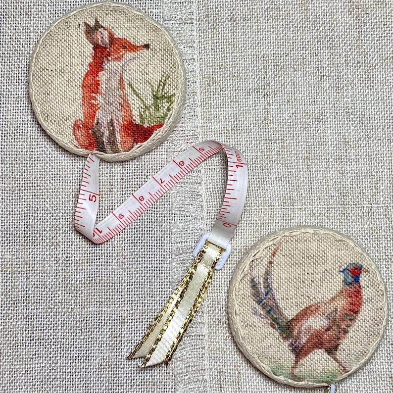 May include: Two round fabric measuring tapes with a fox and a pheasant design. The tapes are beige with brown stitching and feature a white plastic measuring tape with red numbers.