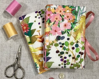 May include: Two fabric covered notebooks with a floral pattern in shades of pink, yellow, and green. The notebooks are stacked on top of each other and are surrounded by sewing supplies, including spools of thread and scissors.