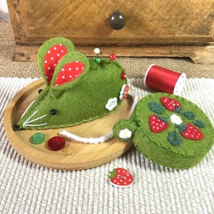 May include: A green felt mouse pincushion with red and white polka dot ears and a white string tail. The pincushion is sitting on a wooden tray with a green felt strawberry pincushion and a spool of red thread.