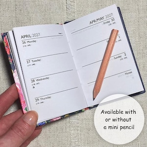Op de afbeelding: Een open zakagenda voor april en mei 2027 met witte pagina's, zwarte tekst en een kaft met bloemenpatroon. Een klein, lichtgekleurd houten potlood ligt diagonaal over de rechterpagina. De tekst luidt Available with or without a mini pencil.
