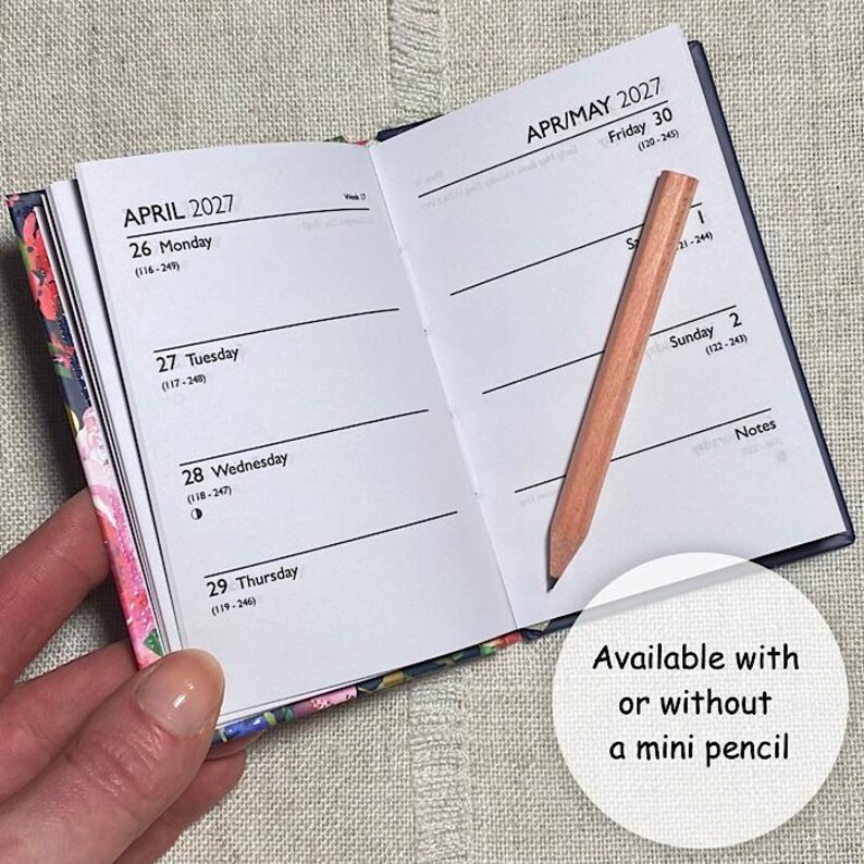 Op de afbeelding: Een open zakagenda voor april en mei 2027 met witte pagina's, zwarte tekst en een kaft met bloemenpatroon. Een klein, lichtgekleurd houten potlood ligt diagonaal over de rechterpagina. De tekst luidt Available with or without a mini pencil.