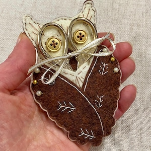 May include: A felt owl-shaped sewing accessory with gold-colored scissors for eyes. The owl is brown and cream-colored, with gold buttons and a white ribbon. The owl's chest is decorated with white stitching and small pearl and gold beads.