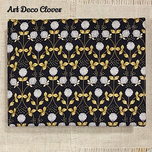 May include: Black fabric with a repeating pattern of white clover flowers and gold leaves. The fabric has a subtle speckled pattern. The text "Art Deco Clovers" is printed in white at the top of the fabric.