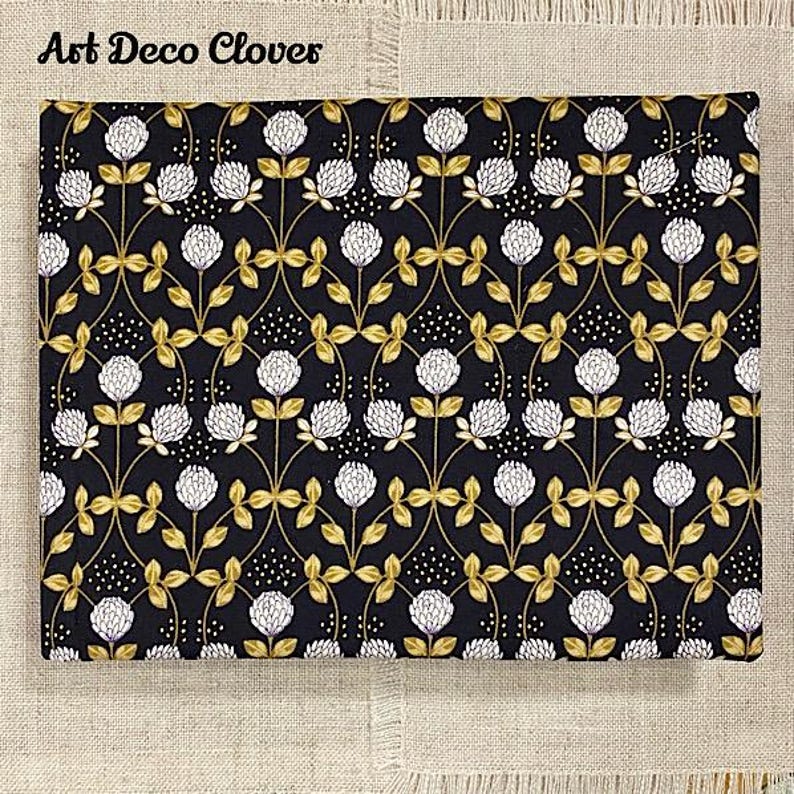 May include: Black fabric with a repeating pattern of white clover flowers and gold leaves. The fabric has a subtle speckled pattern. The text "Art Deco Clovers" is printed in white at the top of the fabric.