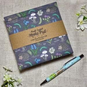 Könnte beinhalten: Ein grauer Notizblock mit einem Waldmuster mit Igeln, Pilzen und Blumen, zusammengehalten von einem braunen Band. Ein Stift mit blauem Blumenmuster liegt neben dem Block. Der Notizblock trägt den Text "Hand Crafted Memo Pad" und "Little Bun Designs UK".