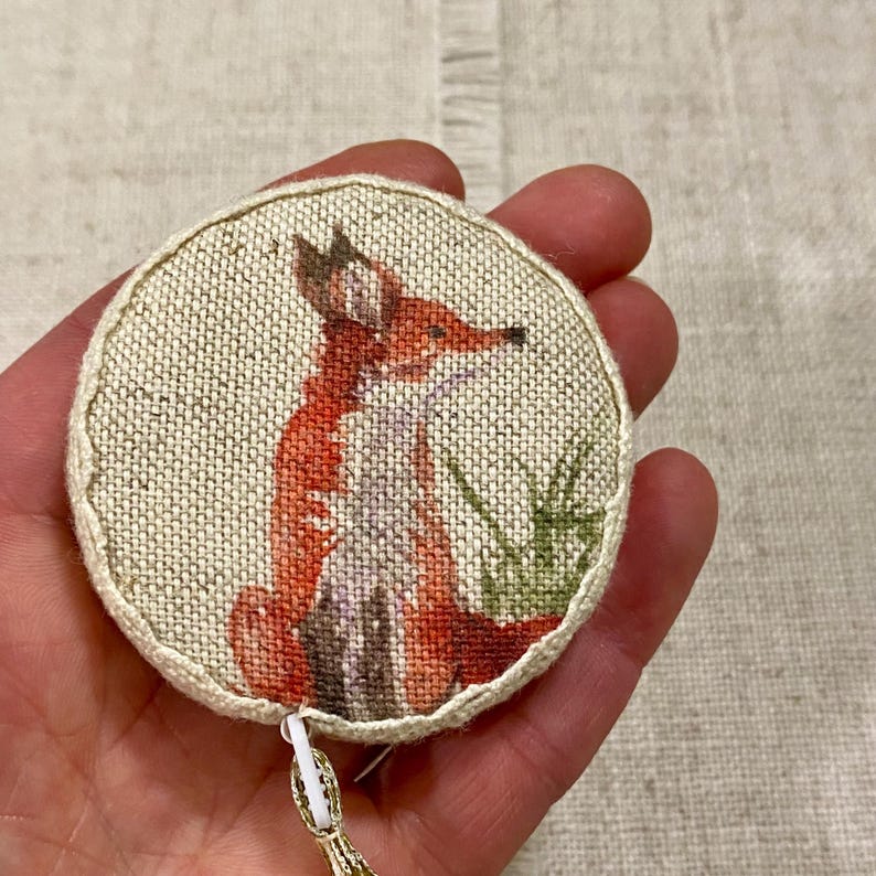 May include: A round, fabric-covered tape measure with a fox design. The fox is orange and white with a brown nose and green grass behind it.