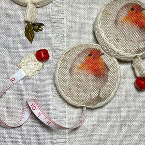 May include: A set of three round fabric measuring tapes with a robin bird design. Each tape has a red bell charm attached. The tapes are on a white linen background.