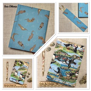 Op de afbeelding: Een blauw dagboek met een zeeotterpatroon, een bijpassende bladwijzer en een tweede dagboek met een kleurrijke print van papegaaiduikers en een kustlandschap. Een pen met patroon en een houten liniaal liggen naast het dagboek.