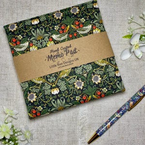 Puede incluir: Un bloc de notas con un diseño decorativo de flores y pájaros en verde, blanco, amarillo y rojo. El bloc está envuelto con una banda de papel marrón que dice "Hand Crafted Memo Pad" y "Little Bun Designs UK". Un bolígrafo a juego está al lado.