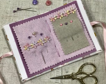 May include: A small pink and white fabric book with two compartments for storing sewing pins and needles. The book is open and has a beige fabric lining with a floral design. There are several pins and needles in each compartment. A pair of gold scissors is lying on a beige fabric surface.