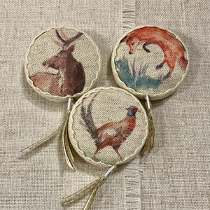 May include: Three round, fabric ornaments with gold tassels. Each ornament features a different animal illustration: a deer, a fox, and a pheasant.