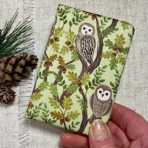 May include: A rectangular fabric case with a repeating pattern of owls on tree branches with green leaves and acorns, set against a light green background. The case is held by a hand.