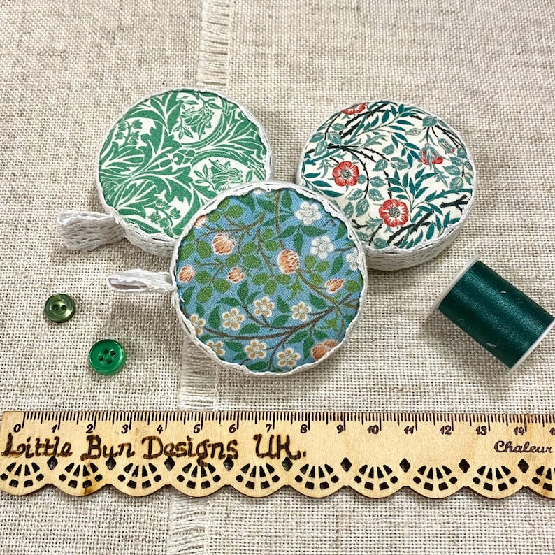 K&ouml;nnte beinhalten: Drei runde Stoffma&szlig;b&auml;nder mit Blumenmustern. Die B&auml;nder sind wei&szlig; mit gr&uuml;nen, blauen und roten Blumendesigns. Die B&auml;nder liegen auf einer beigefarbenen Stoffoberfl&auml;che mit einem Holzlineal, auf dem "Little Bun Designs UK" steht, und einer Spule gr&uuml;nen Fadens.
