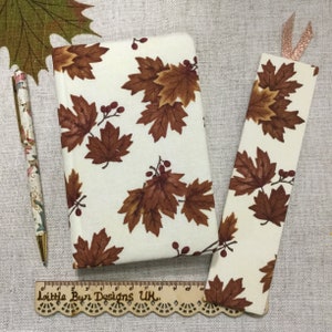 Pode incluir: Um conjunto de diário, marcador e caneta de cor creme. O diário e o marcador apresentam um padrão repetido de folhas de bordo castanhas e bagas vermelhas. A caneta tem um design floral. Há também uma régua de madeira com o texto "Little Byn Designs UK".