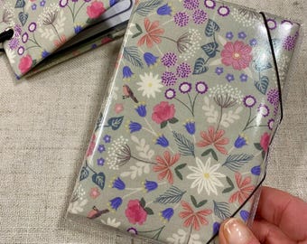 Op de afbeelding: Notitieboekhoes met bloemenpatroon, met roze, paarse en witte bloemen en kleine vogels op een grijze achtergrond. Het heeft een transparante plastic overlay en een zwarte elastische band.