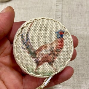 May include: A round, cream-colored fabric measuring tape with a colorful pheasant design. The tape is edged with a white stitch.