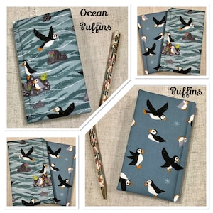Op de afbeelding: Een met stof bekleed notitieboek met een patroon van papegaaiduikers op een blauwe oceaanachtergrond met golvende witte lijnen en rotsen, vergezeld van een bijpassende pen. De tekst Ocean Puffins en Puffins staat op de afbeelding.