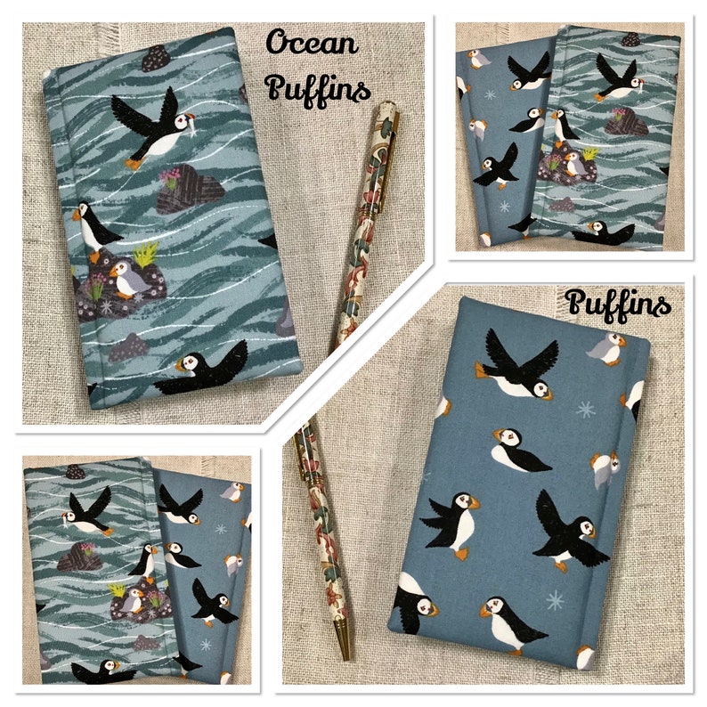 Op de afbeelding: Een met stof bekleed notitieboek met een patroon van papegaaiduikers op een blauwe oceaanachtergrond met golvende witte lijnen en rotsen, vergezeld van een bijpassende pen. De tekst Ocean Puffins en Puffins staat op de afbeelding.