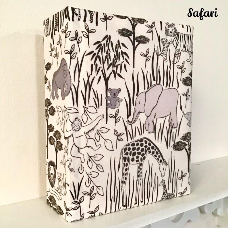 May include: A white fabric with a black and brown safari animal print. The print includes a giraffe, elephant, monkey, gorilla, and tiger. The fabric has the word "Safari" printed in the top right corner.