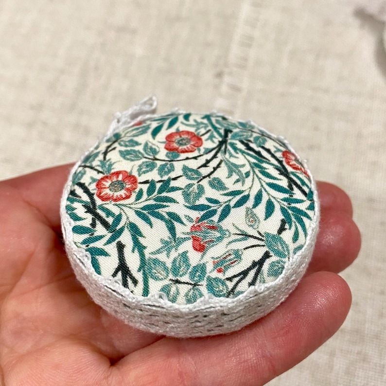 K&ouml;nnte beinhalten: Ein rundes, wei&szlig;es, geh&auml;keltes Nadelkissen mit einem Blumenmuster in Gr&uuml;n- und Rott&ouml;nen. Der Stoff zeigt ein sich wiederholendes Muster aus Bl&auml;ttern und Bl&uuml;ten.