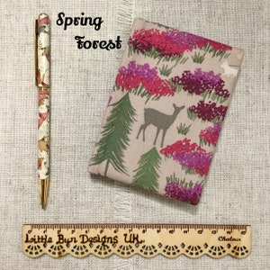 May include: A pen and a fabric case with a forest theme. The pen has a gold tip and a cream-coloured body with a floral pattern. The case features a deer, trees, and flowers in pink and purple. A wooden ruler is also present.