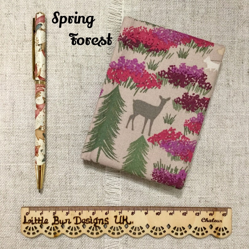 May include: A pen and a fabric case with a forest theme. The pen has a gold tip and a cream-coloured body with a floral pattern. The case features a deer, trees, and flowers in pink and purple. A wooden ruler is also present.