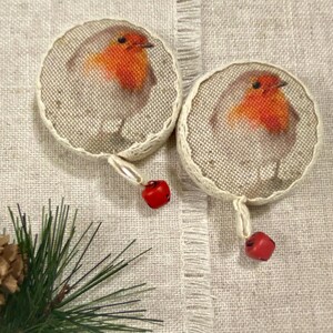 May include: Two round, white, fabric ornaments with a robin bird design. Each ornament has a red bell attached to a silver cord.