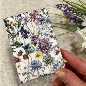 May include: A white fabric gift wrap with a floral pattern featuring various flowers in shades of purple, pink, yellow, and green. The flowers are arranged in a random pattern on a white background.