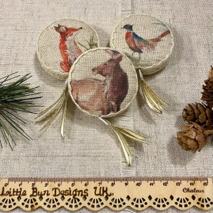 May include: Three round burlap ornaments with gold tassels. Each ornament features a different woodland animal: a fox, a deer, and a pheasant. The ornaments are on a beige fabric background.