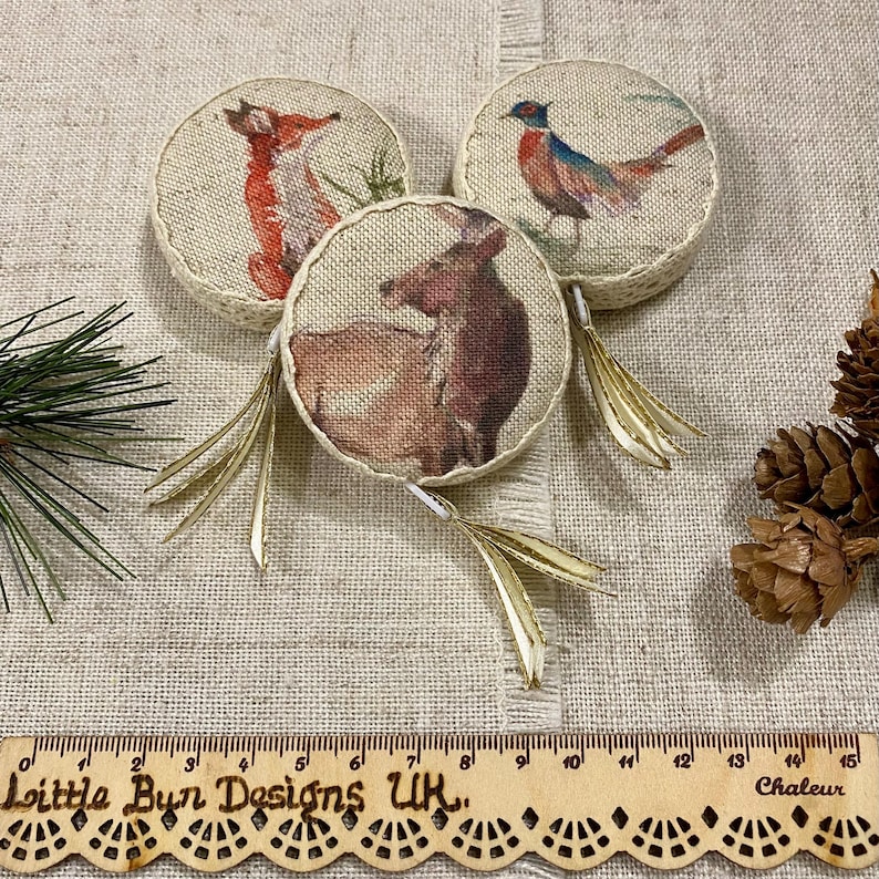 May include: Three round burlap ornaments with gold tassels. Each ornament features a different woodland animal: a fox, a deer, and a pheasant. The ornaments are on a beige fabric background.