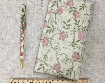 Op de afbeelding: Een stoffen etui met Jasmine Ivory patroon en een bijpassende balpen met goudkleurige details. Een houten liniaal onderaan toont dat de voorwerpen ongeveer 15 centimeter lang zijn. De stof heeft een bloemenprint met roze en witte bloemen en groene bladeren op een ivoorkleurige achtergrond.