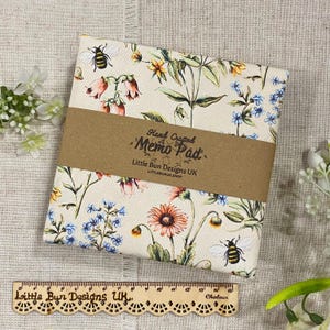 Könnte beinhalten: Ein floraler Notizblock mit cremefarbenem Hintergrund, mit bunten Blumen und Bienen. Der Block ist mit einem braunen Papierband mit der Aufschrift "Hand Crafted Memo Pad" umwickelt. Ein Holzlineal befindet sich darunter.