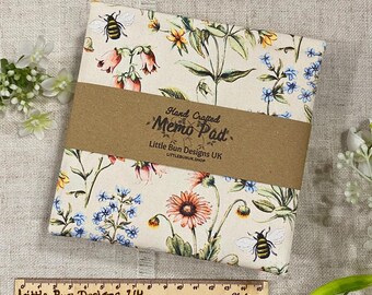 Op de afbeelding: Een bloemenmemopad met een crèmekleurige achtergrond, met kleurrijke bloemen en bijen. Het pad is verpakt met een bruine papieren band met de tekst "Hand Crafted Memo Pad". Een houten liniaal staat eronder.