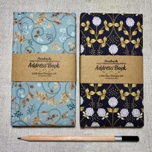 May include: Two address books with brown paper bands labeled Handmade Using Traditional Bookbinding Methods Address Book Little Bun Designs UK. One book has a light blue cover with brown leaves and acorns, the other a dark blue cover with white flowers and gold stems. A wooden pencil lies below.